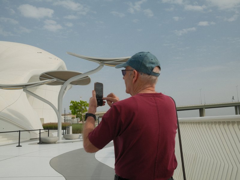 An elderly man in a maroon shirt and blue cap takes a photo with his smartphone against a modern architectural backdrop.
