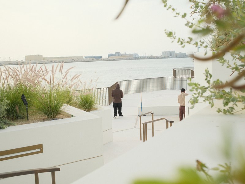 Two people standing by a waterfront with greenery and a city skyline in the background.