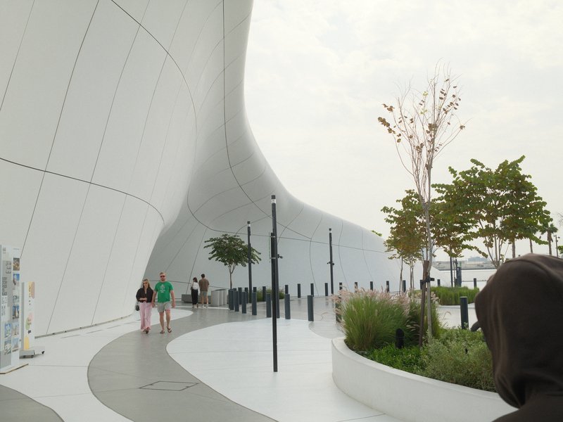 A modern architectural structure with a curved white facade, people walking, and greenery nearby.