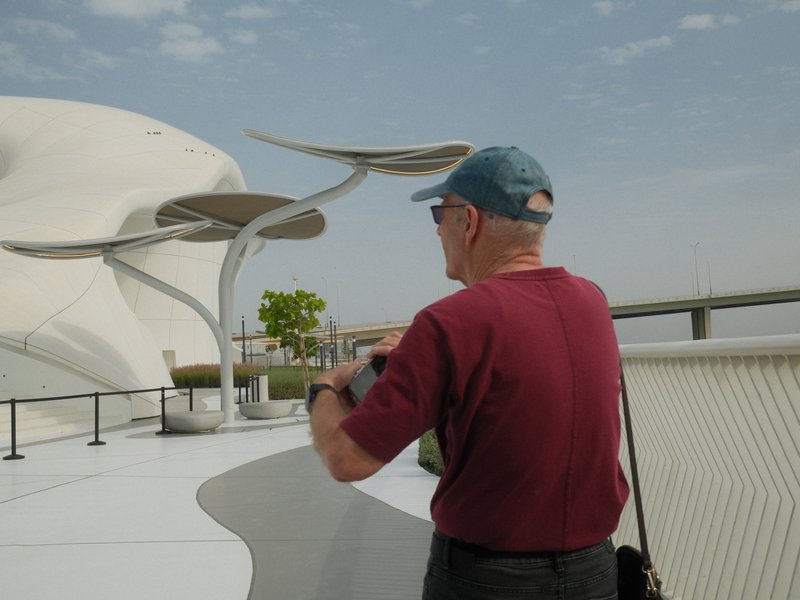 An elderly man in a red shirt and blue cap stands on a modern architectural walkway, taking a photo.