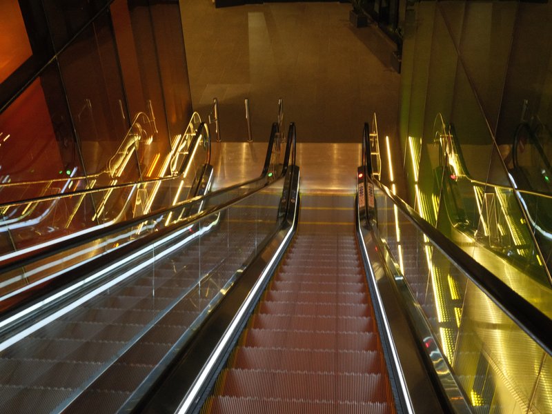 A view down an escalator with illuminated sides and colorful reflections on the walls.