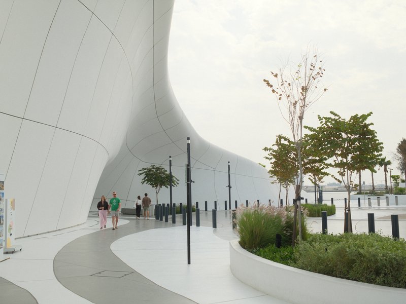 A modern architectural structure with curved white walls, trees, and people walking along a path.