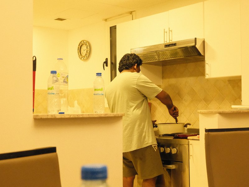 A person cooking in a modern kitchen with light-colored walls and a clock on the wall.