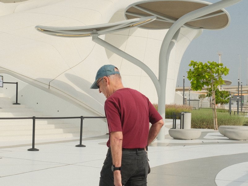 An elderly man in a maroon shirt and cap walks away from a modern architectural structure.