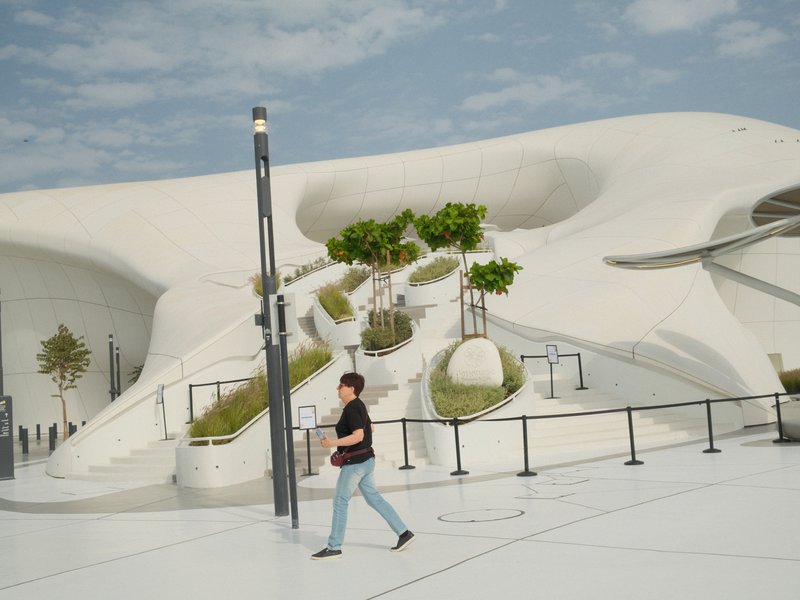 A person walking near a modern, white architectural structure with greenery and blue sky.