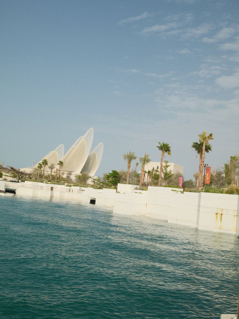 A modern architectural structure near a calm body of water, with palm trees and a clear blue sky.