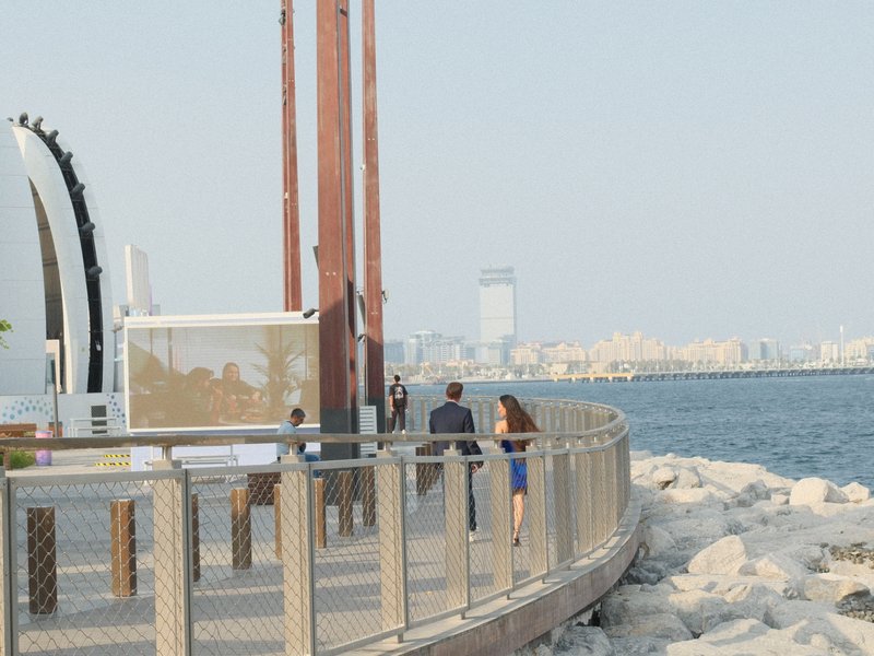 Couple walking along a waterfront path with city skyline in the background on a clear day.