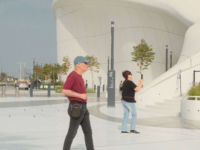 Two people walking near a modern architectural structure with trees and a cloudy sky.