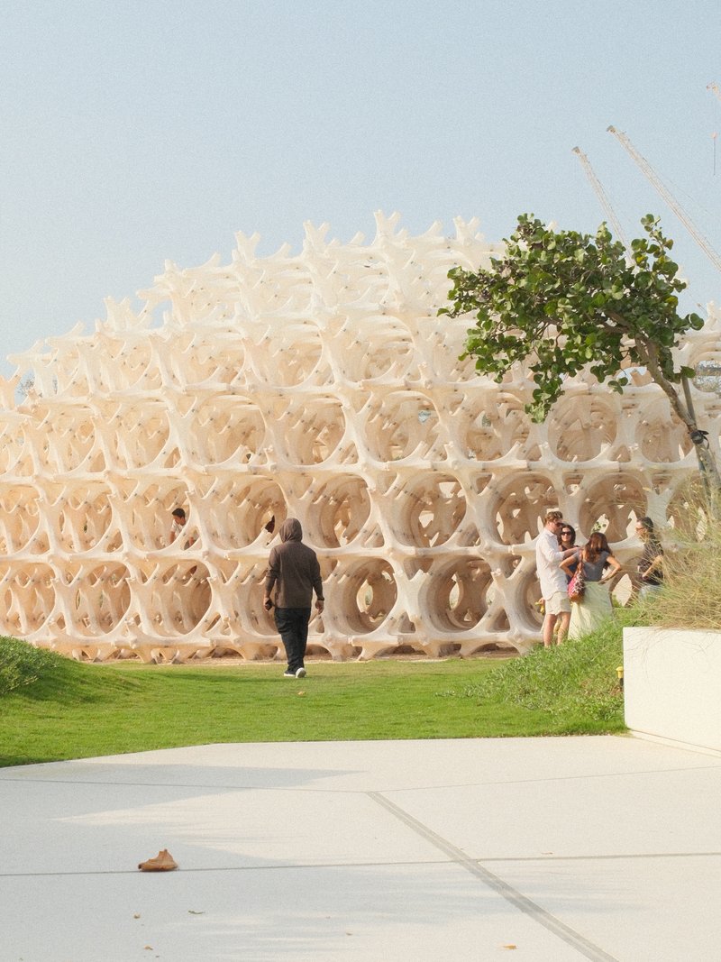 A modern architectural structure resembling a honeycomb, with people walking nearby in a green space.