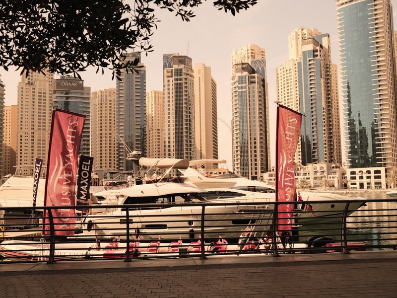 A luxury yacht docked by a waterfront with tall skyscrapers in the background and flags fluttering.