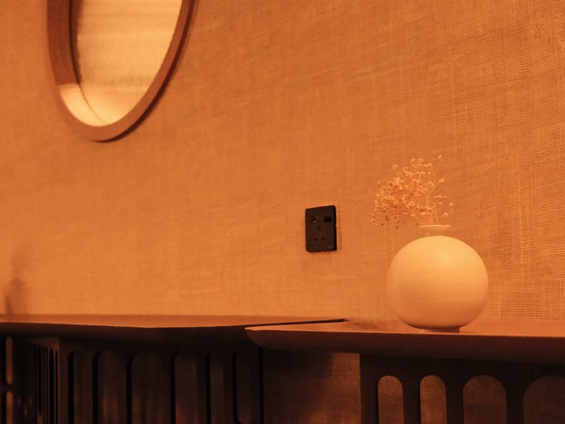 A minimalist interior scene featuring a round vase with dried flowers on a wooden shelf against a textured wall.