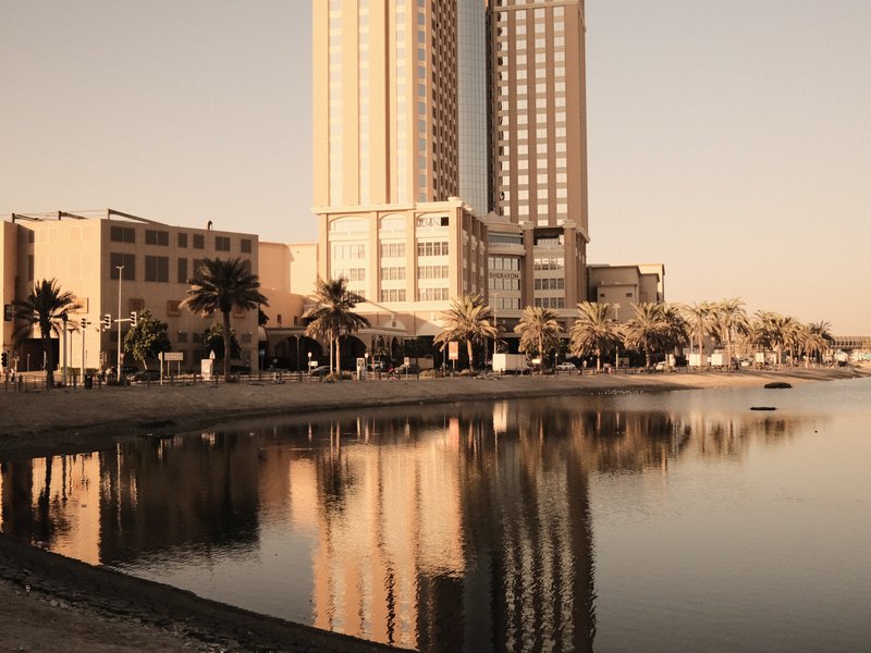 A tall building reflecting in calm water during golden hour, with palm trees lining the shore.