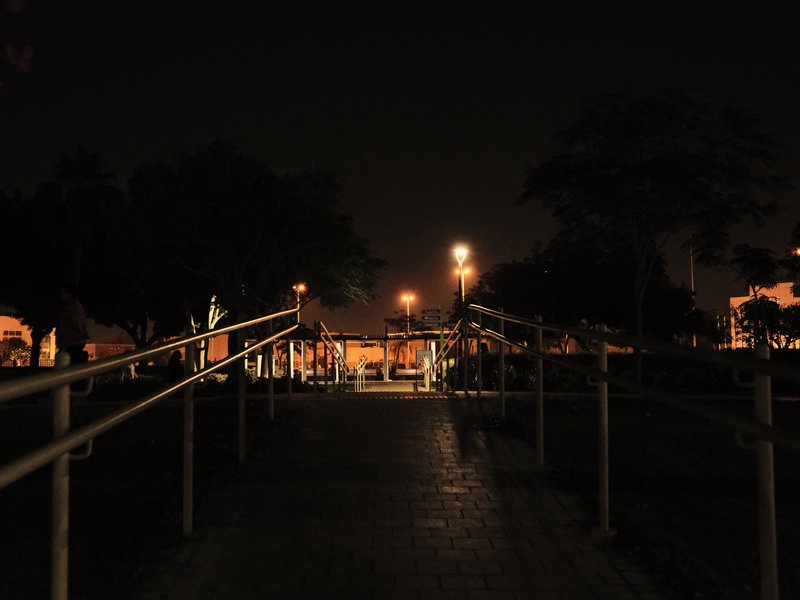 A dimly lit pathway with railings leading towards a distant light source in a park at night.