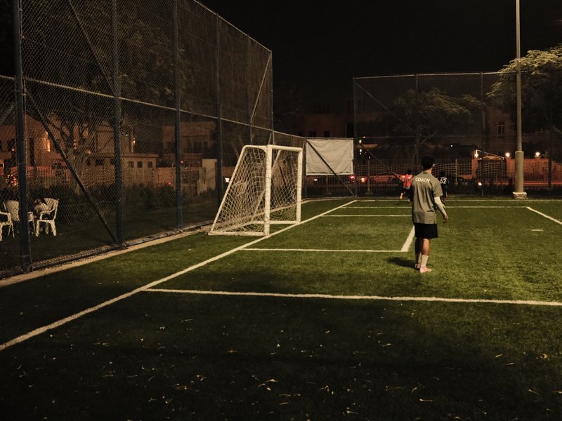 A person walking towards a soccer goal on a dimly lit field at night, surrounded by a fence.