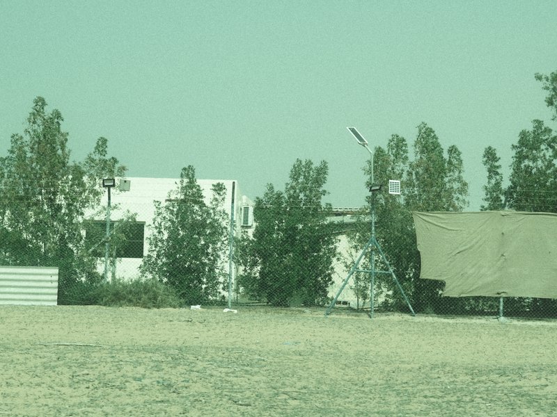 A distant view of a white building partially obscured by trees, with a tent in the foreground on sandy ground.