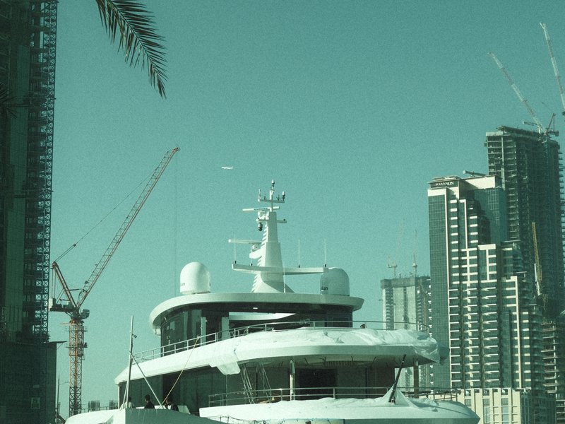 A luxury yacht docked near modern skyscrapers with construction cranes in the background.