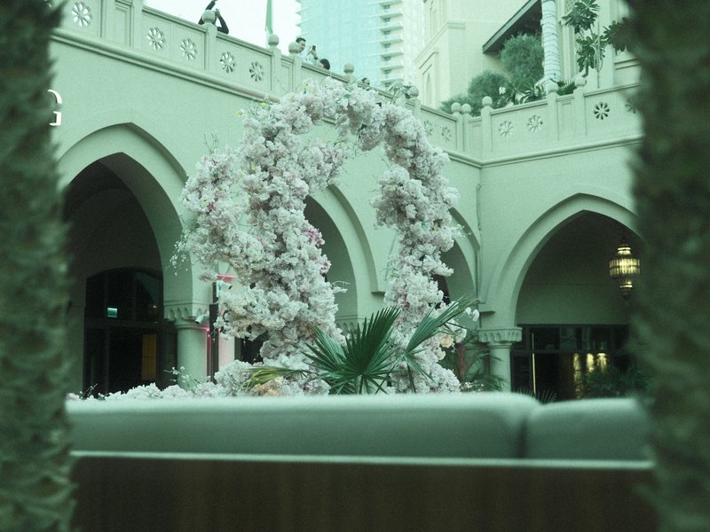 A floral arch made of pink blossoms in an indoor courtyard with arches and greenery.