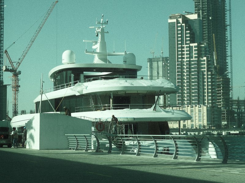 A large yacht docked near modern skyscrapers with construction cranes in the background.