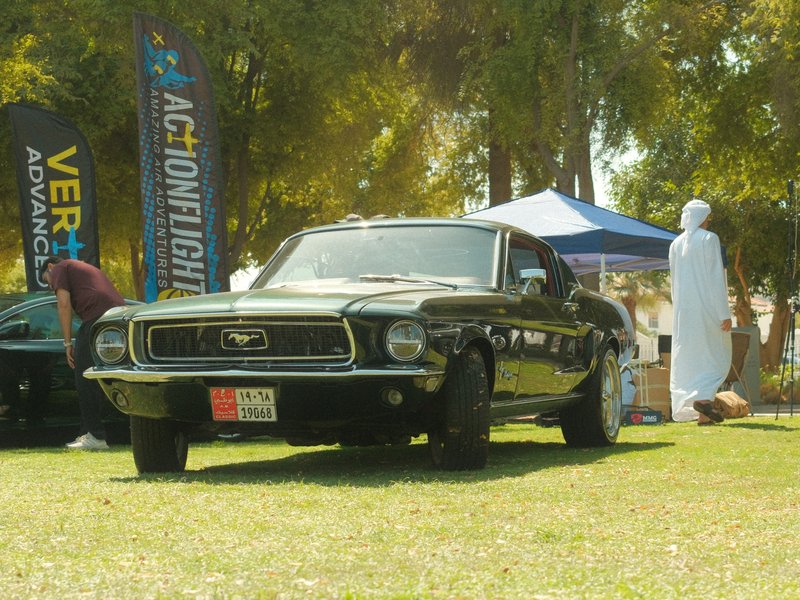 A classic black Mustang parked on grass at an outdoor event with tents in the background.