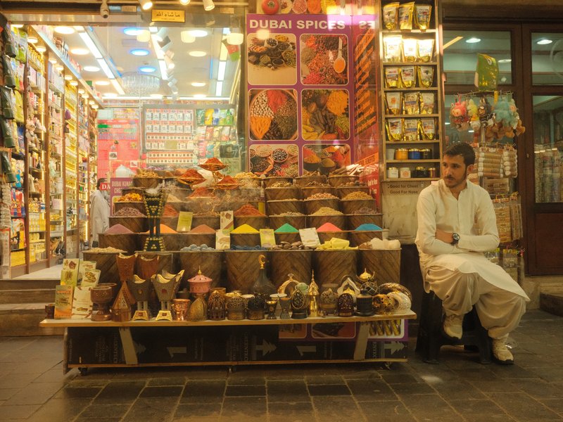 A vendor sitting beside a colorful display of spices and goods in a market setting.