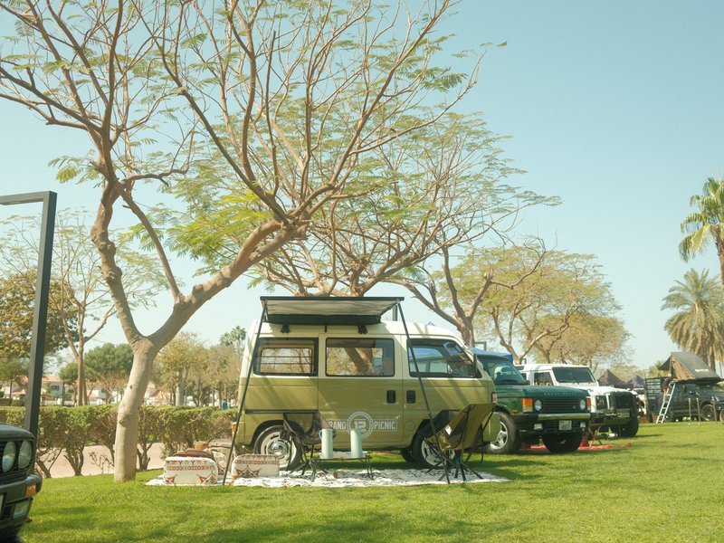 A green camper van parked under trees in a sunny outdoor setting with other vehicles in the background.