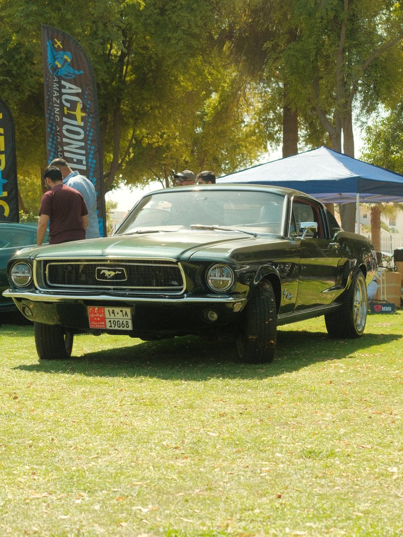 A classic black Mustang car parked on grass with people and tents in the background.