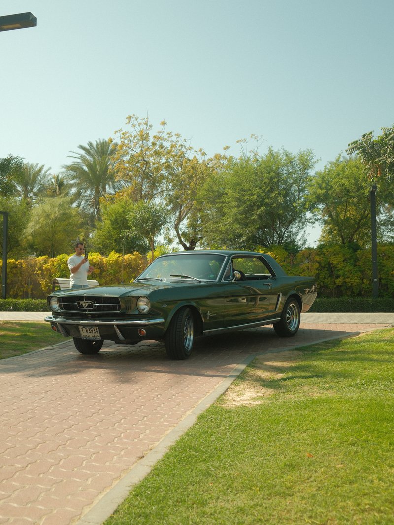 A classic green Mustang parked on a sunny pathway surrounded by lush greenery and palm trees.