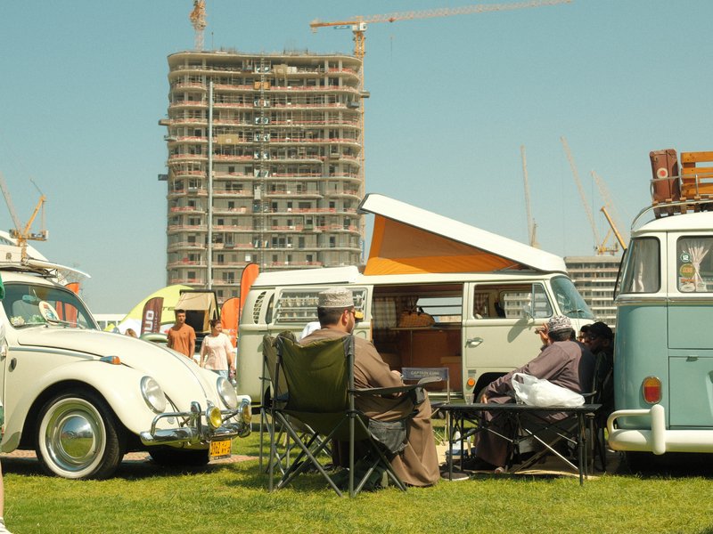 Two men sitting in camping chairs near vintage cars at a sunny outdoor event with a construction site in the background.