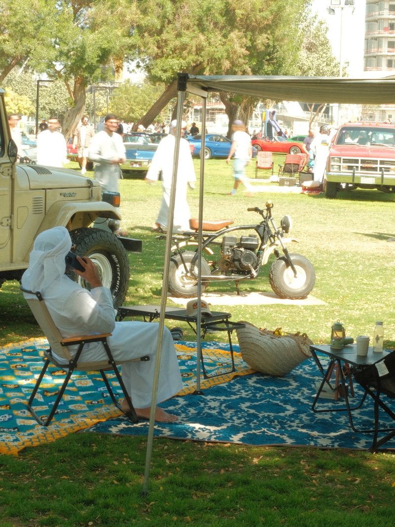 A man in traditional attire sitting on a chair under a canopy in a park, with vehicles and people in the background.