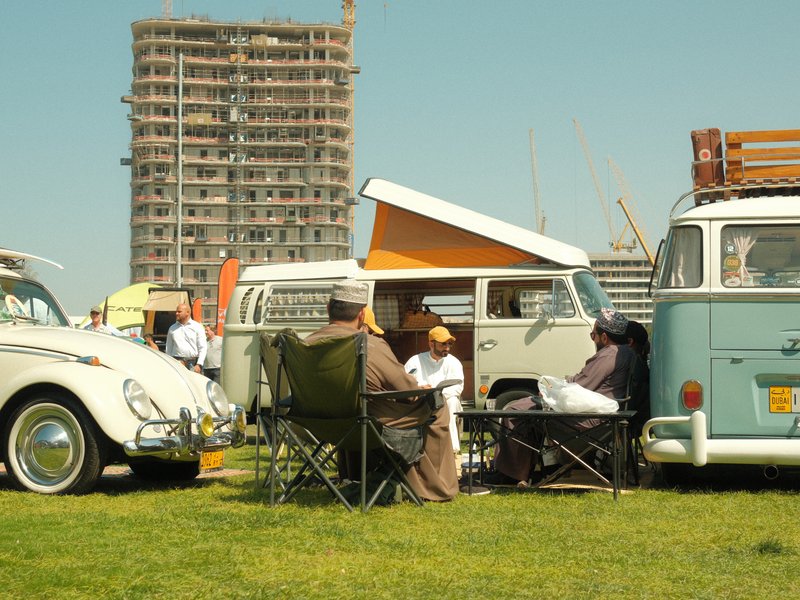 A group of people relaxing in chairs near vintage cars at a sunny outdoor event with a building under construction in the background.