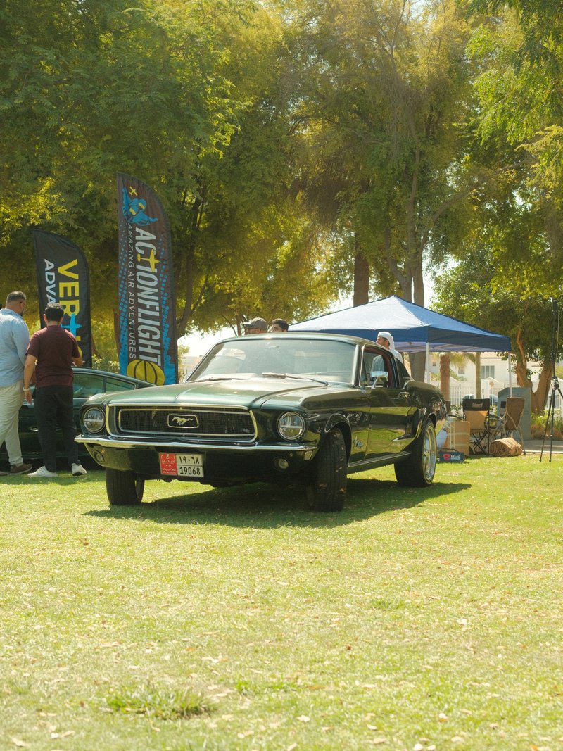 A classic black Mustang parked on grass with people and tents in the background.