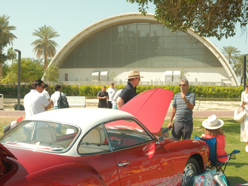 A classic red car with its hood open in a park, surrounded by people and palm trees.