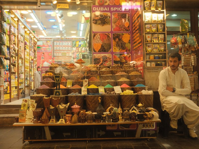 A man sitting outside a colorful shop filled with spices and decorative items at night.