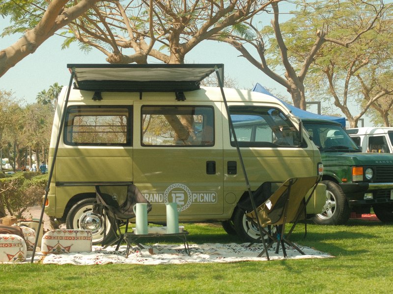 A vintage camper van parked under trees with camping chairs set up in front on a grassy area.