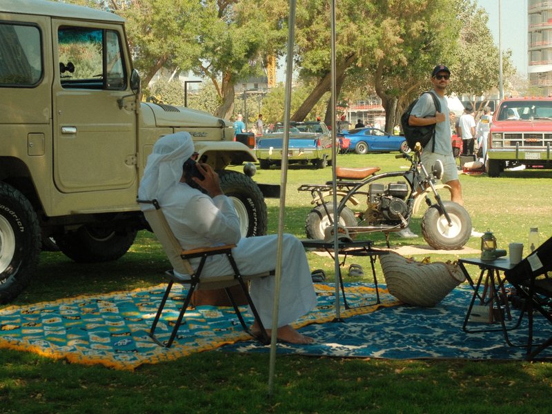 A man in traditional attire sitting under a tent with a vintage vehicle nearby in a park setting.