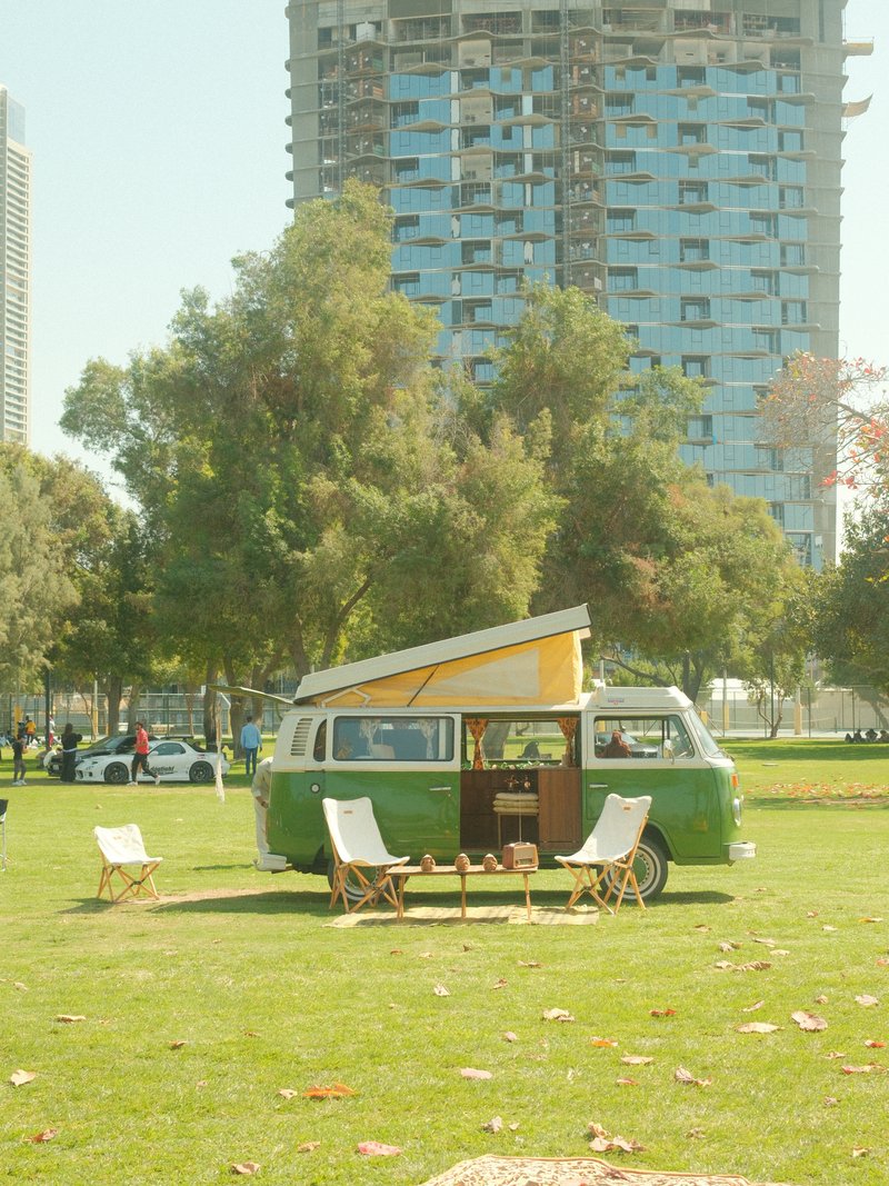 A vintage green camper van parked on a grassy area with modern buildings in the background.