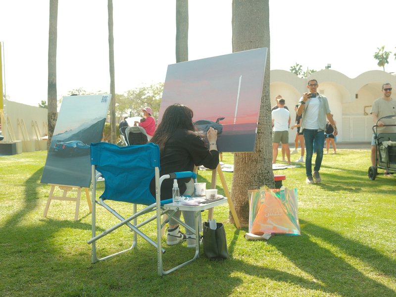 An artist painting outdoors with two canvases, surrounded by people and palm trees in a sunny park.