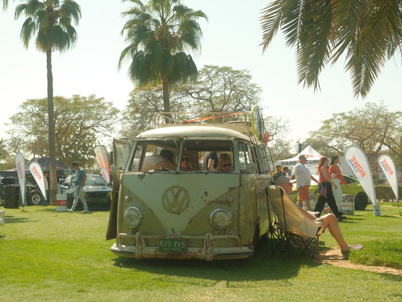 A vintage Volkswagen bus parked on a grassy area with palm trees and people in the background.