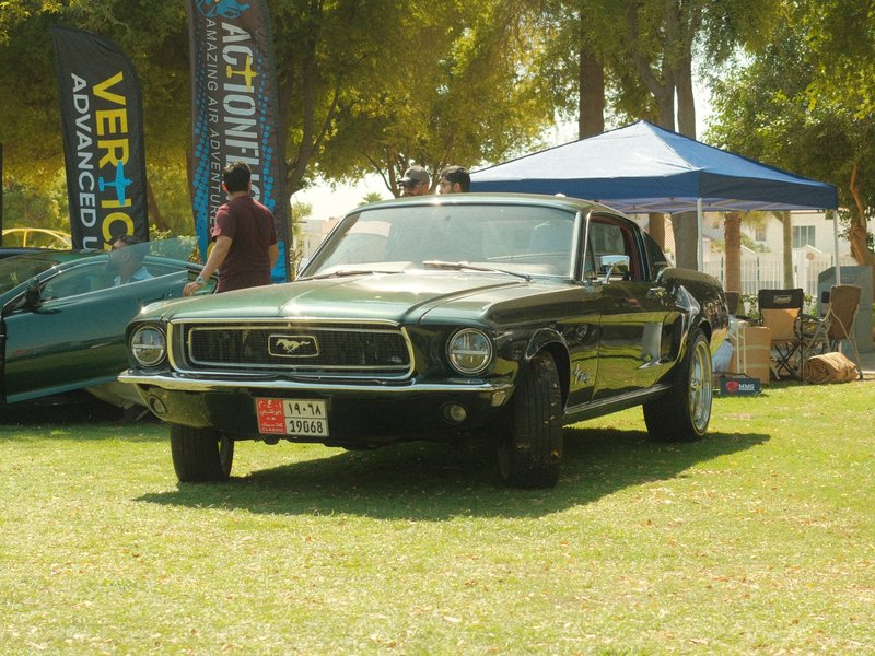 A classic black Mustang car parked on grass at an outdoor event with tents in the background.