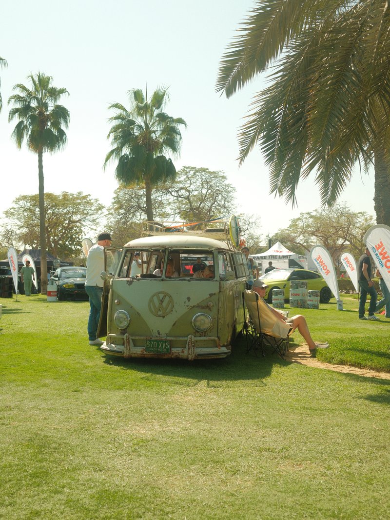 A vintage Volkswagen van parked on grass with palm trees in the background and people nearby.