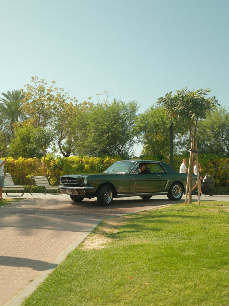 A classic green car parked on a sunny path with people walking in a park.