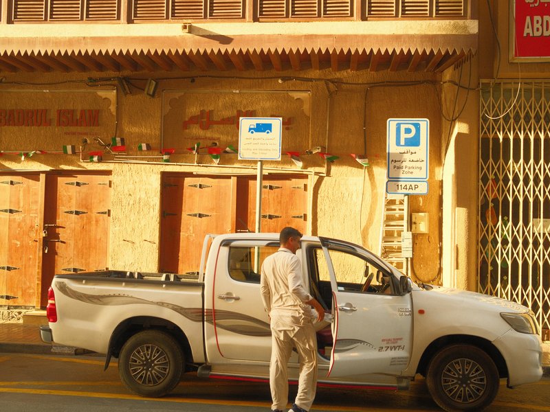 A man in light clothing stands next to a white pickup truck in a sunlit urban setting.