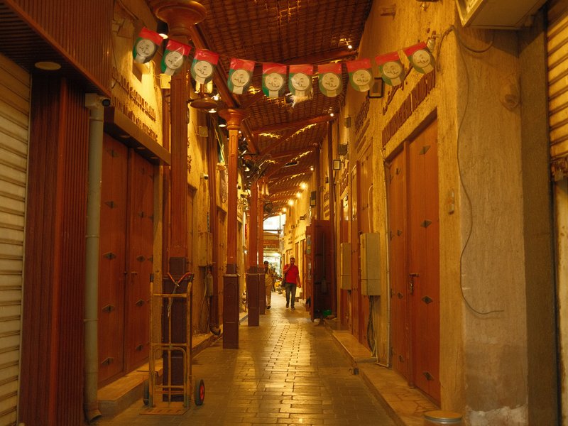 Narrow alleyway in a market with wooden doors and decorative lights overhead.