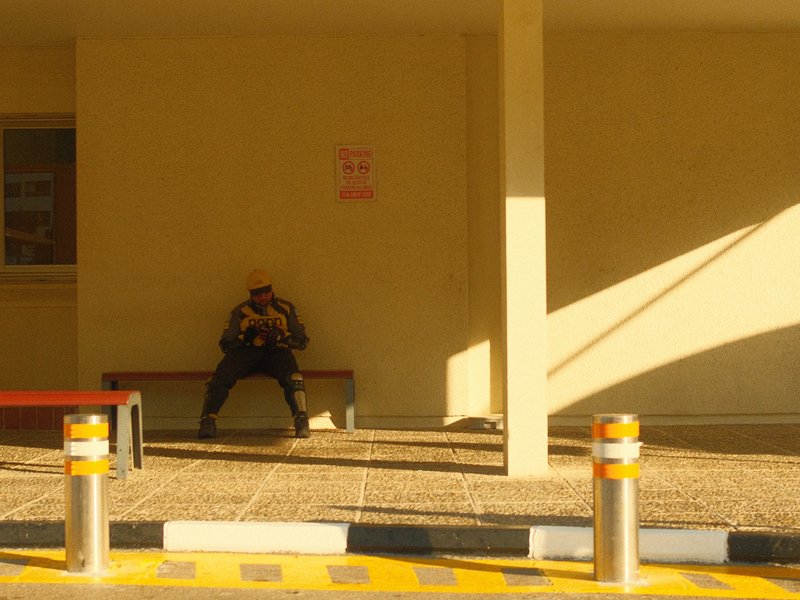 A solitary figure sitting on a bench in a brightly lit, minimalistic space with shadows.