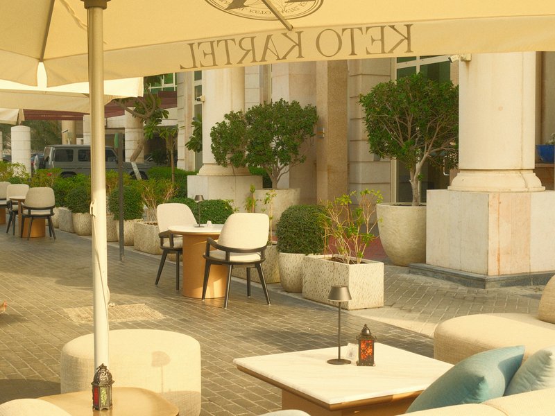 Outdoor café scene with tables and chairs under an umbrella, surrounded by greenery.