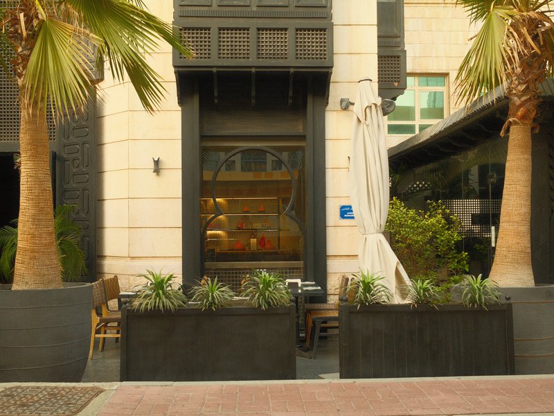 A modern restaurant entrance with palm trees and planters, featuring a cozy outdoor seating area.
