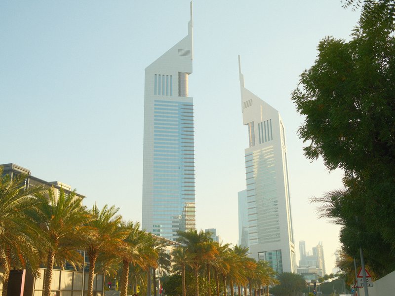 Two tall modern skyscrapers with unique architecture against a clear sky, framed by palm trees.