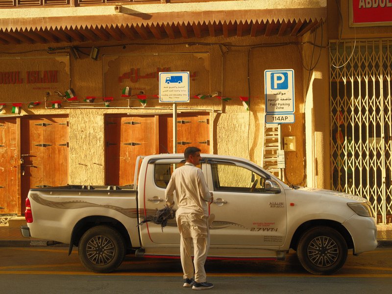 A person in light clothing stands next to a white pickup truck in an urban setting with wooden doors.