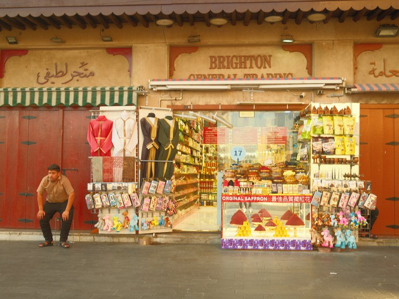 A colorful storefront with various goods displayed, a man sitting nearby, and vibrant signage.
