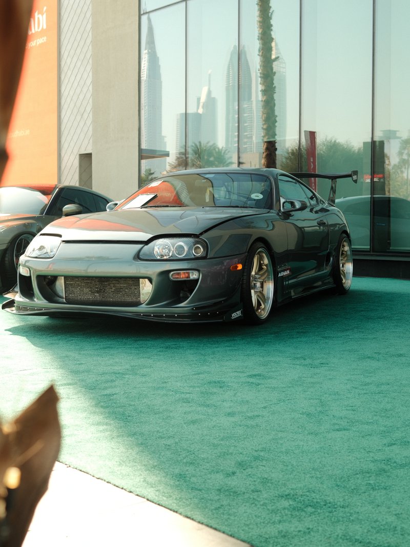 A modified sports car parked on green carpet with skyscrapers in the background.